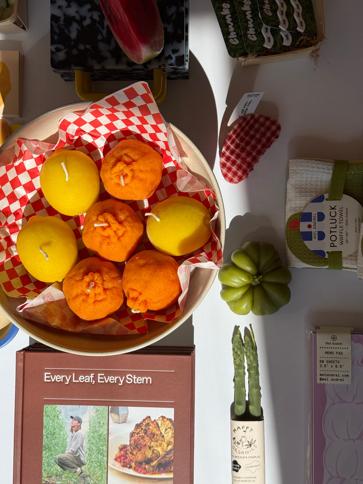 Flatlay of shop table with a bowl of citrus candles, tea towel, cookbook, tomato candle, asparagus candle, and hair clip in the direct sunlight.